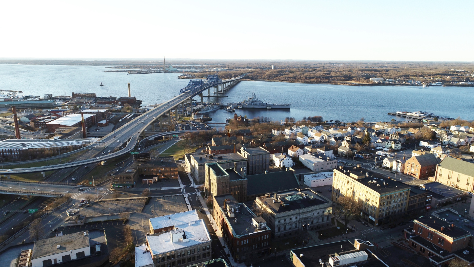 Aerial view of the Braga Bridge crossing the Taunton River in Fall River, Massachusetts, with the USS Massachusetts battleship and Battleship Cove visible at the waterfront.
